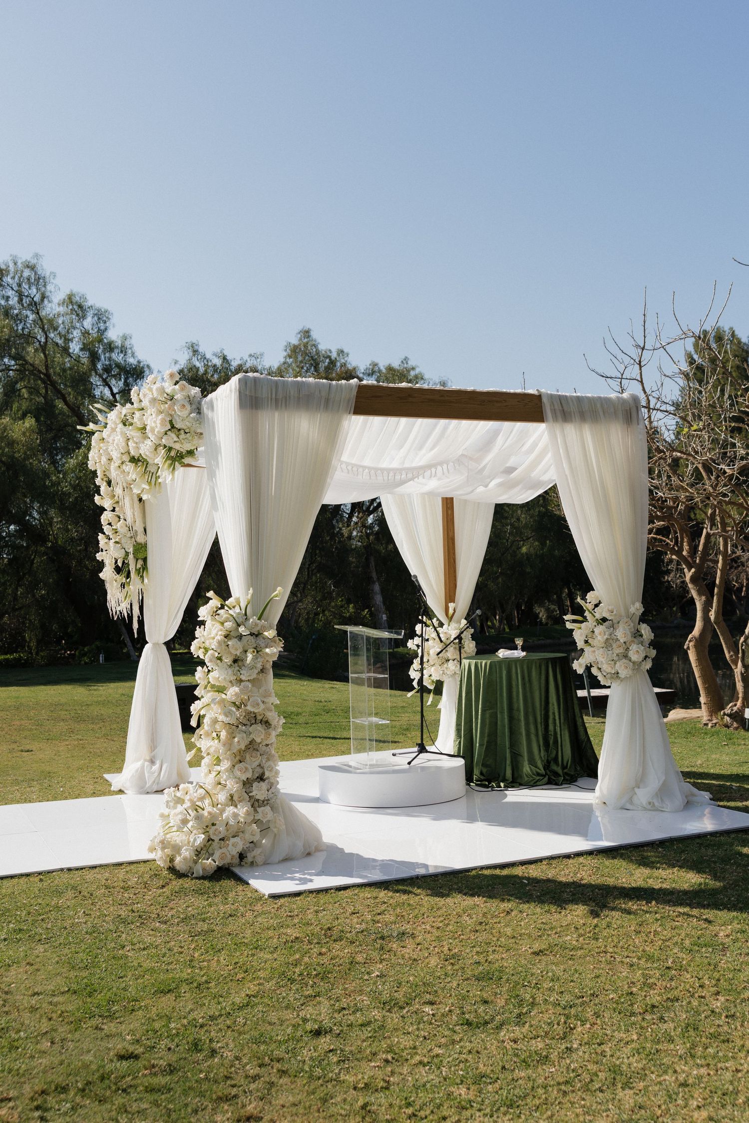 Elegant outdoor wedding chuppah with white draping and floral arrangements on green lawn.
