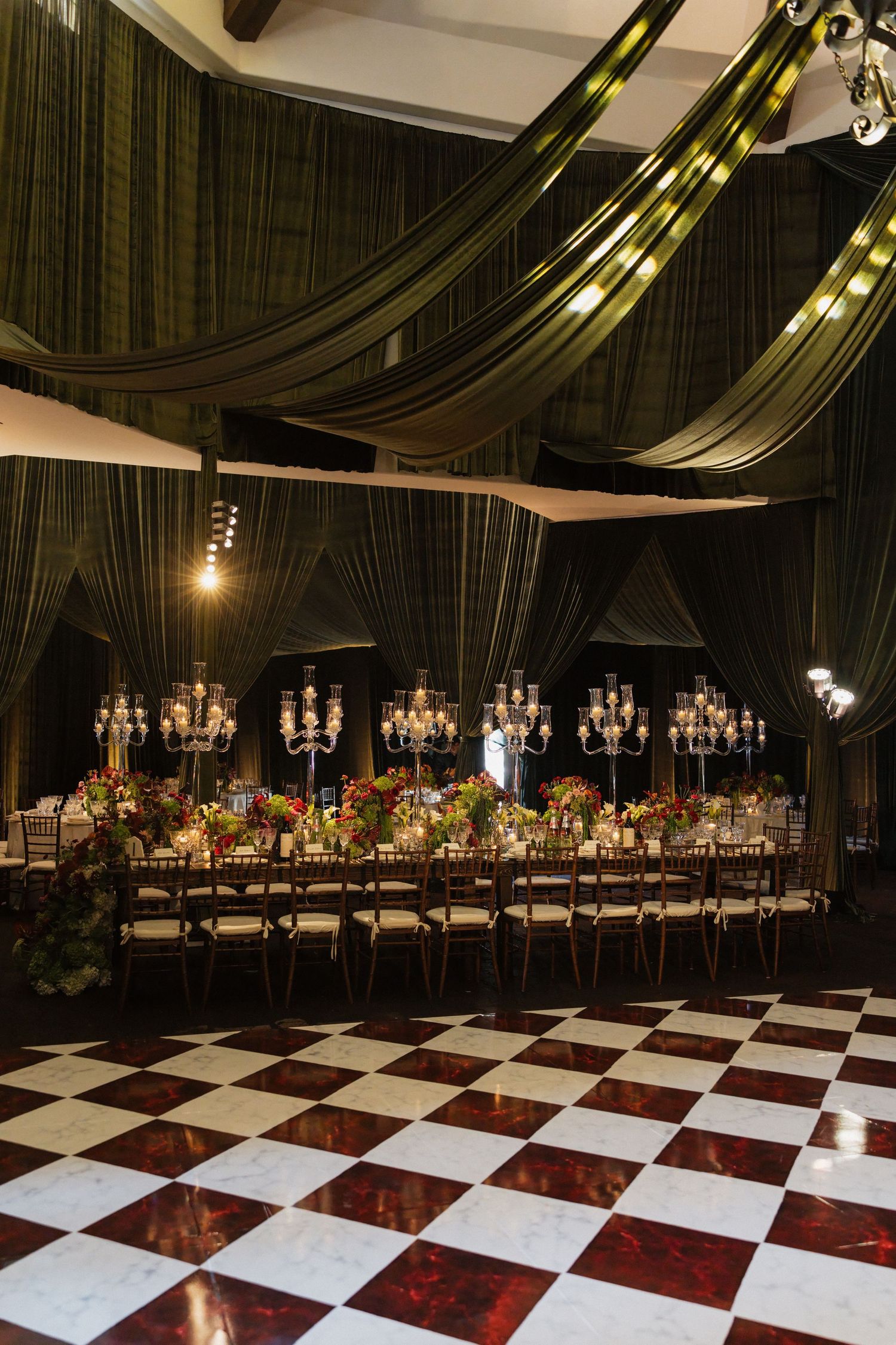 Elegant wedding reception hall with dark green draping, crystal candelabras, long banquet table, and black-and-white checkered dance floor.