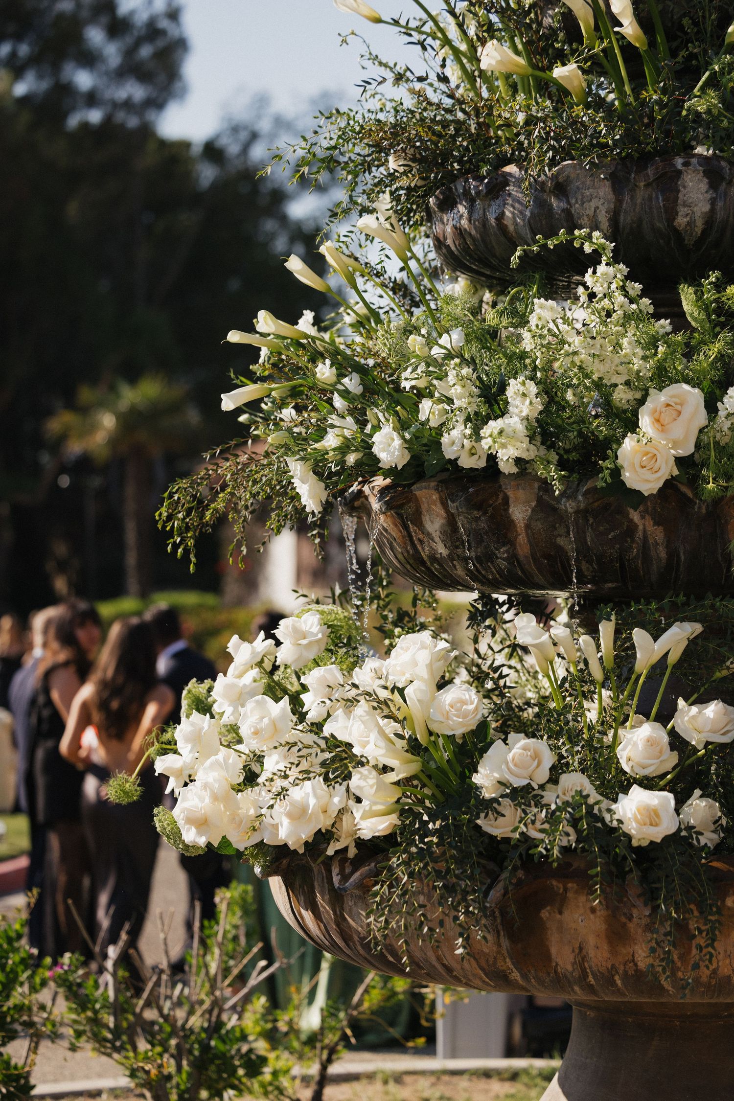 Close-up of a tiered stone fountain decorated with white roses and lilies at an outdoor wedding.
