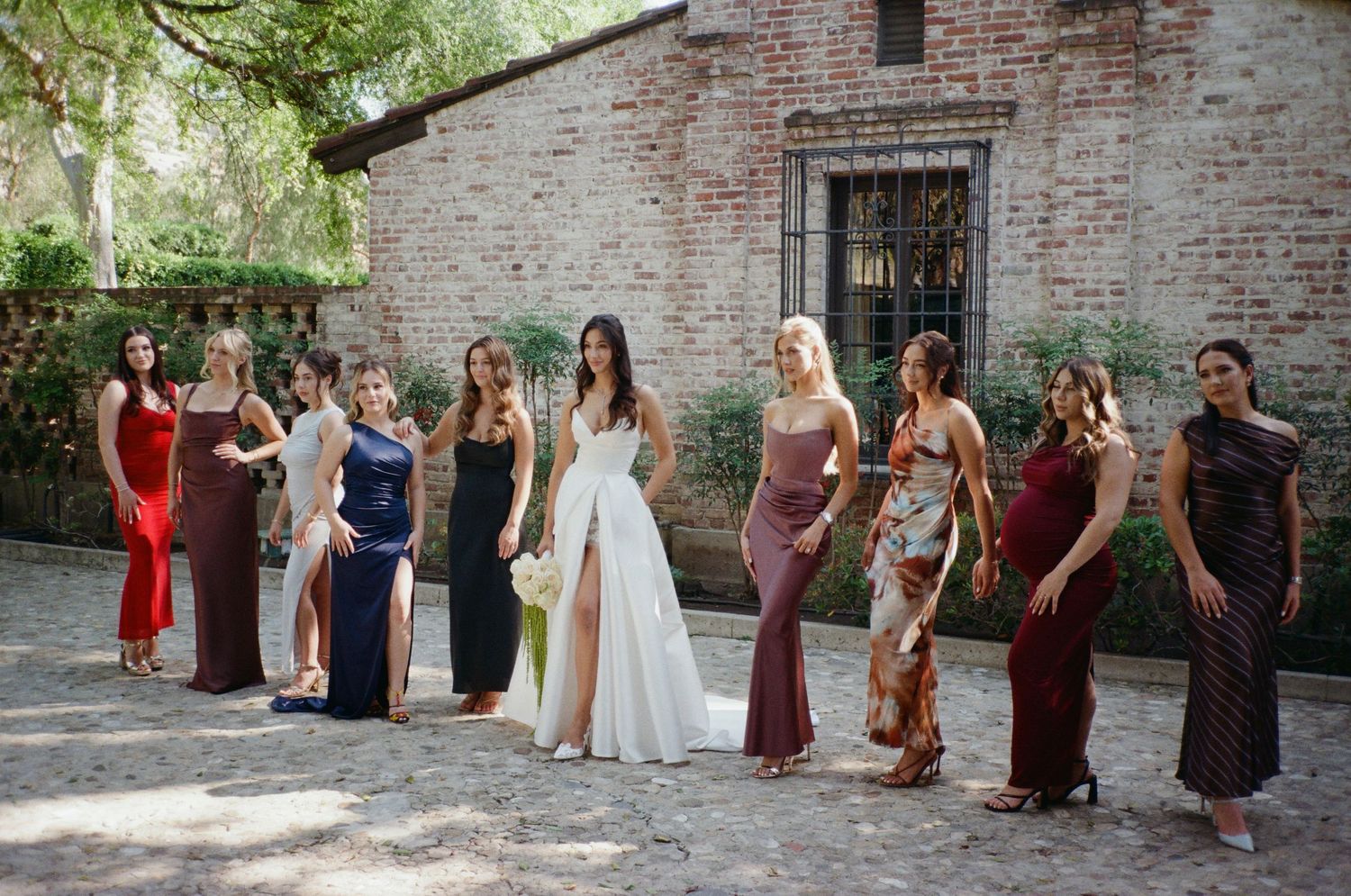 Bride in white gown poses with bridesmaids in colorful dresses outside rustic brick building.