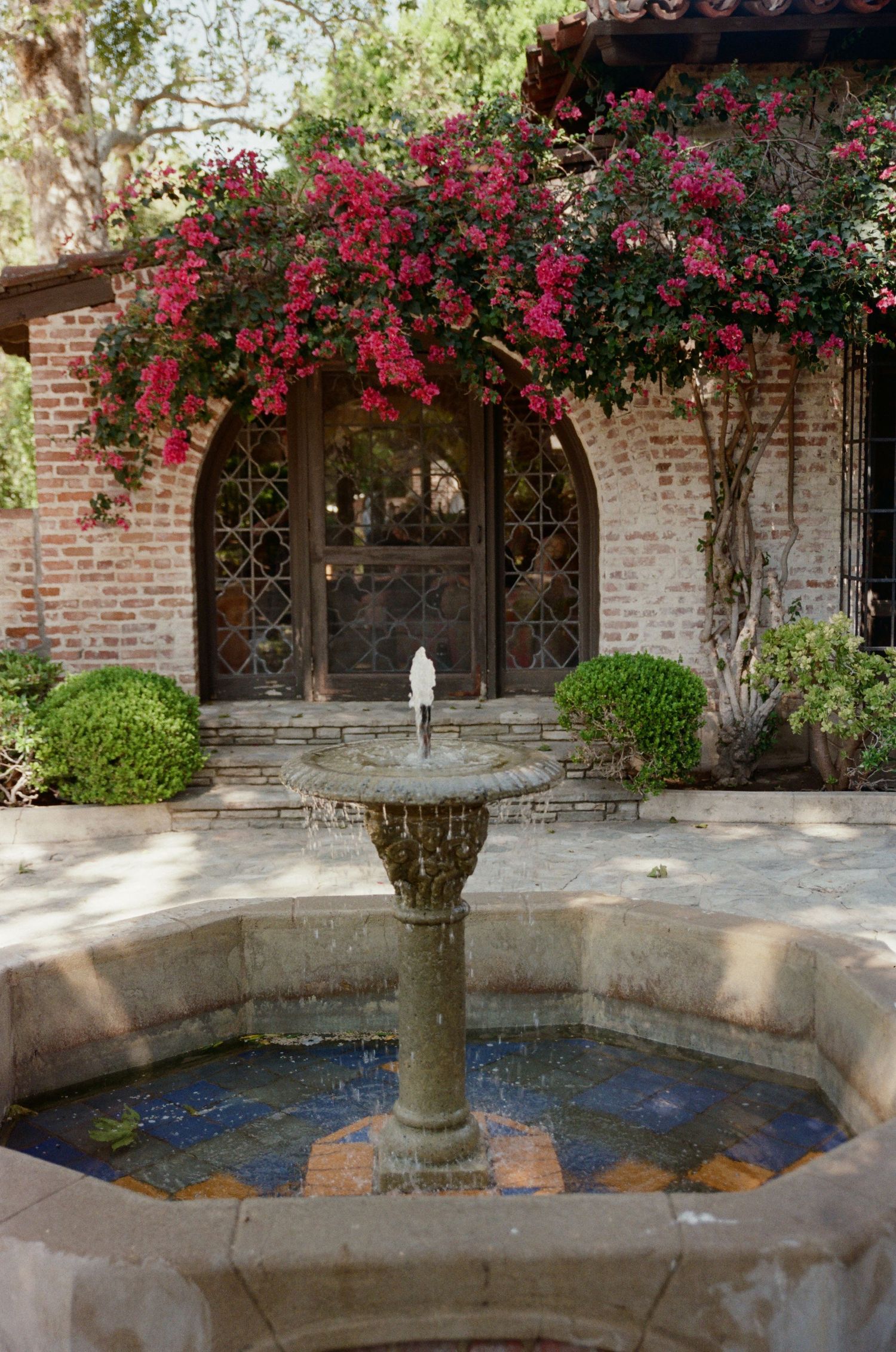 Stone fountain with water feature in a Spanish-style courtyard with bougainvillea blooms over brick archway.