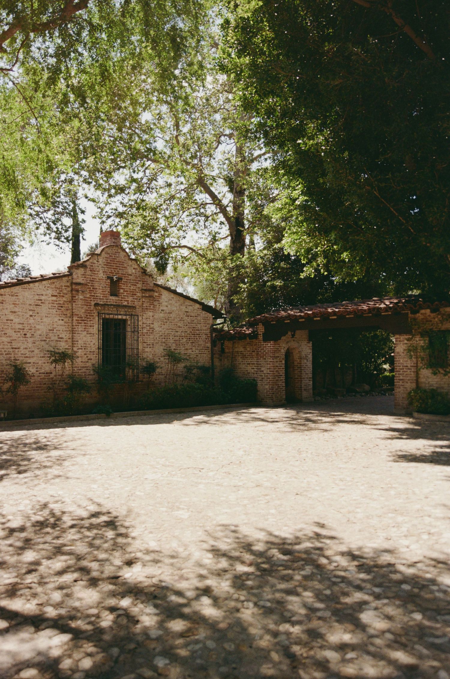 Sunlit brick courtyard with historic adobe buildings and lush green trees casting shadows on gravel ground.