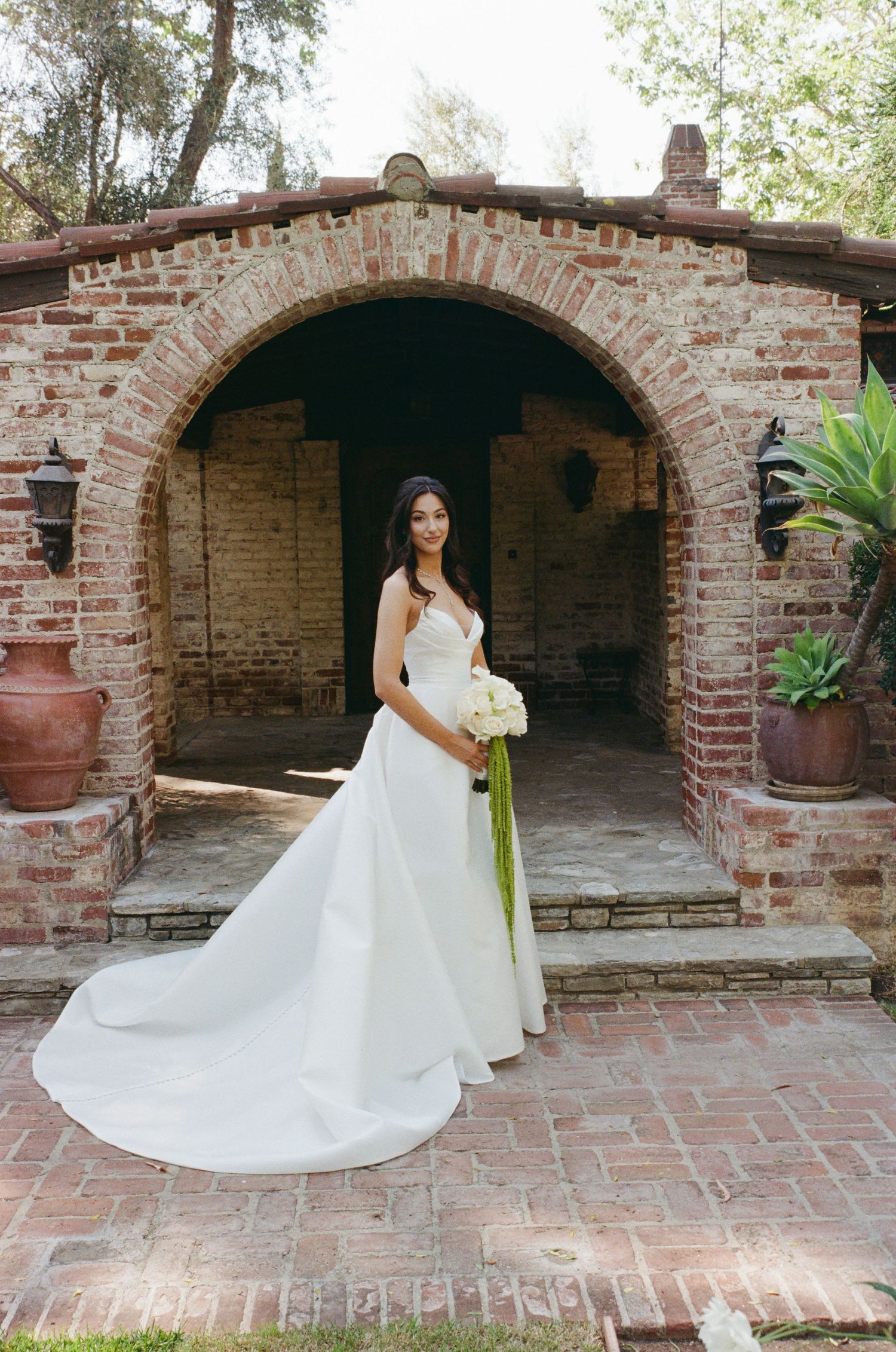 Bride in elegant white gown with long train holds white bouquet under brick archway at outdoor wedding venue.