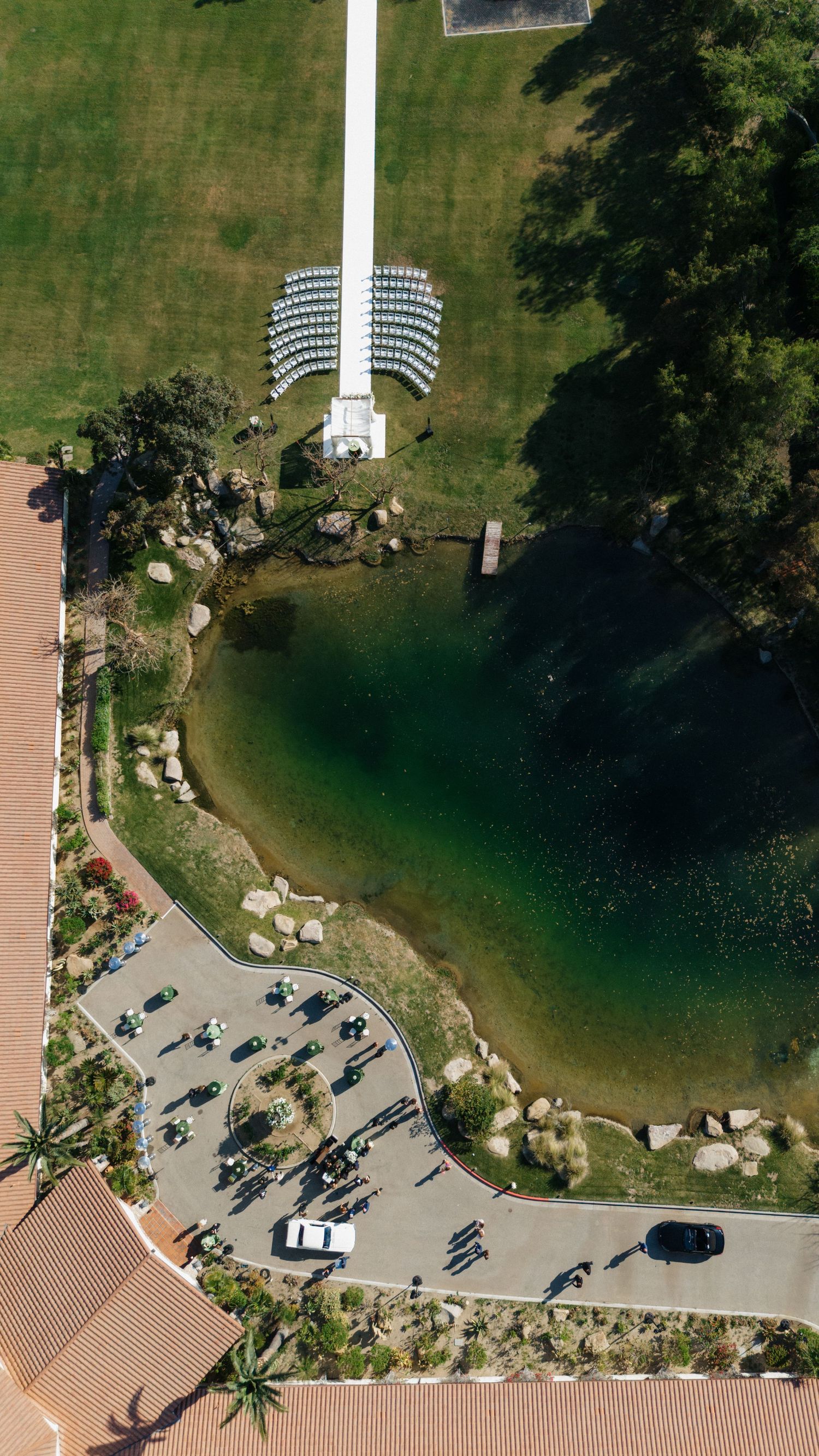 Aerial view of a scenic venue with a pond, green lawn, seating arrangement, and parking area surrounded by trees.