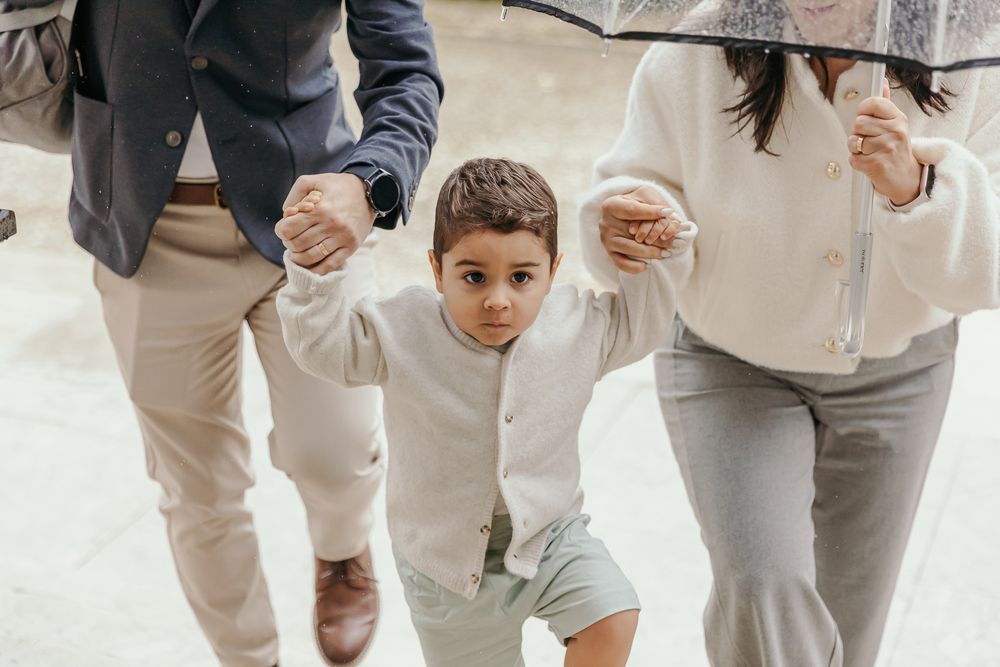 Young boy in white outfit holds parents hands while walking in rain under umbrella.