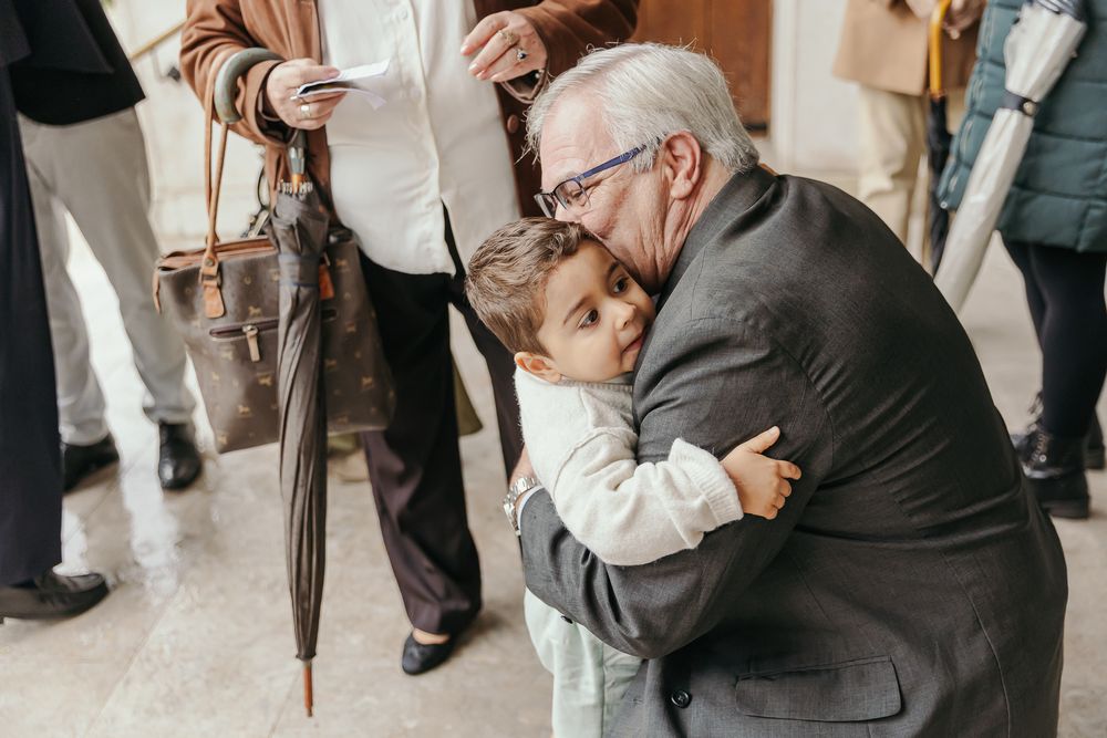 Elderly man hugging a young child warmly at a family gathering, with people holding umbrellas in the background.