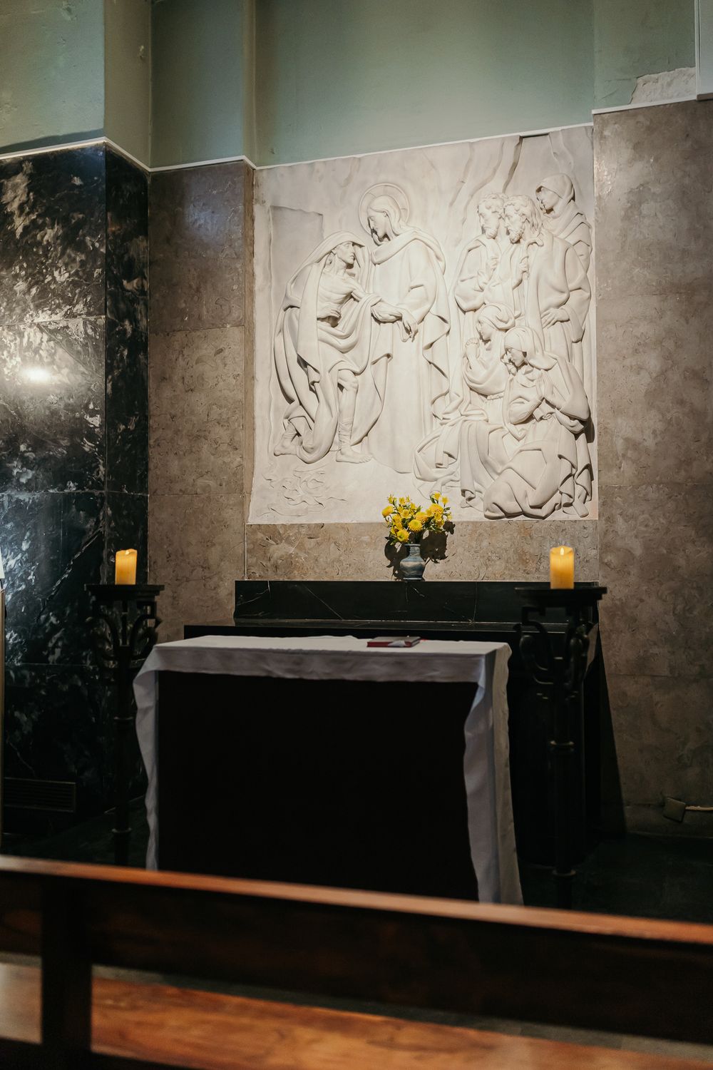 Stone relief sculpture above a candlelit altar with yellow flowers in a serene church chapel interior.