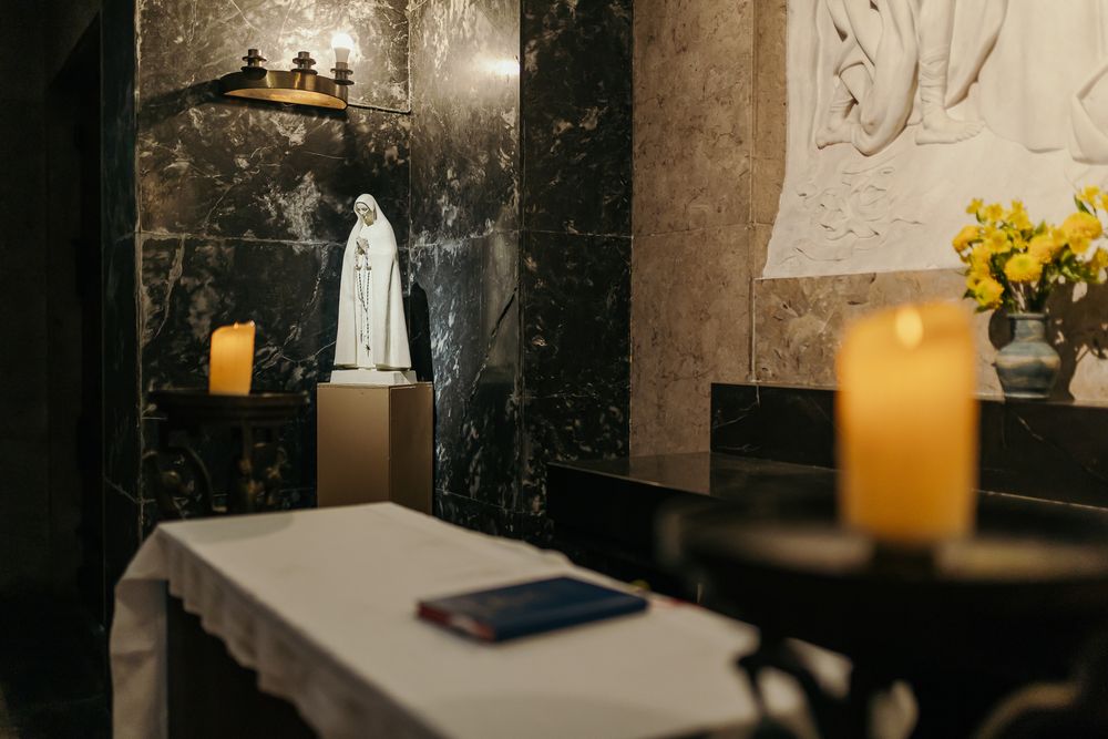 A dimly lit chapel with a Our Lady of Fatima statue, lit candles, and yellow flowers on a dark marble altar.