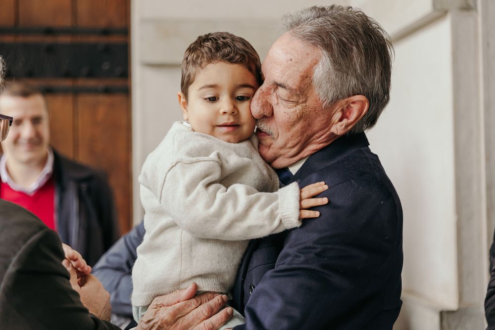 Elderly man tenderly kissing a young toddler boy on the cheek while holding him in his arms indoors.