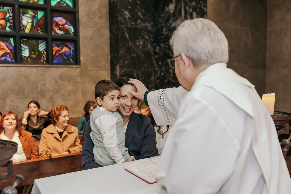 Priest blessing a young child held by a parent during a church ceremony with congregation seated in background.