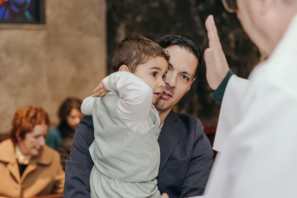 Man holding a young child during a religious ceremony in a church, with a priest raising his hand in blessing.