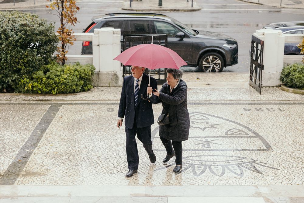 Two people walking in rain sharing a red umbrella on a wet plaza with cars in background.