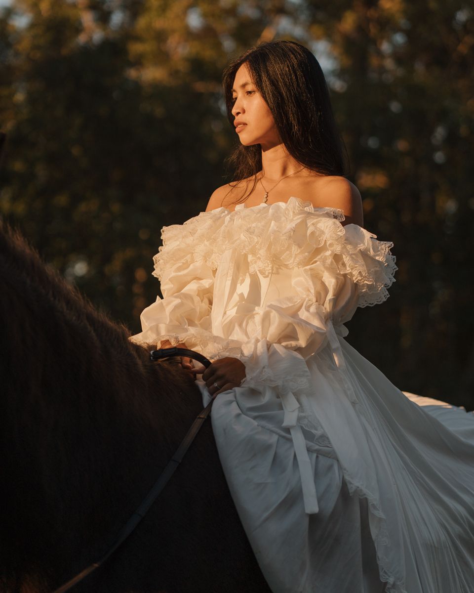 A young woman in a white ruffled off-shoulder gown rides a dark horse at golden hour photoshoot with a horse in sydney