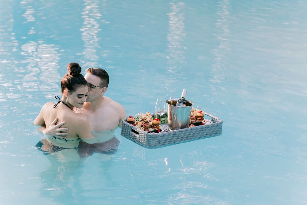 Couple embracing in a pool beside a floating tray with drinks and snacks on a sunny day.