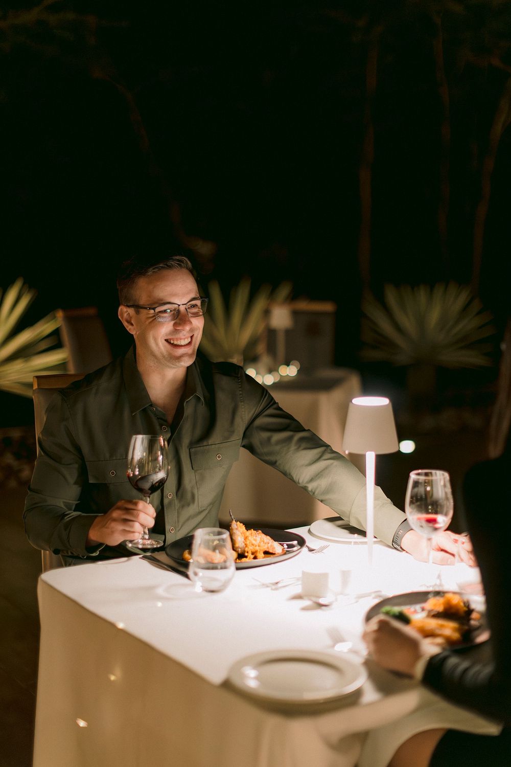 Man smiling and holding red wine at an elegant outdoor dinner table lit by a small lamp at night.
