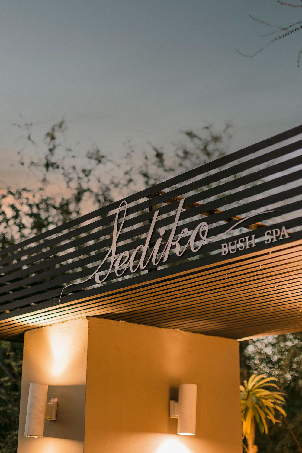 Sediko Bush Spa entrance sign illuminated by warm wall lights at dusk with trees in background.