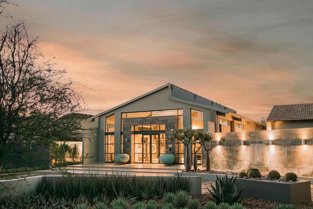 Elegant modern building exterior glowing at dusk with warm lights, surrounded by lush landscaping and dramatic sunset sky.