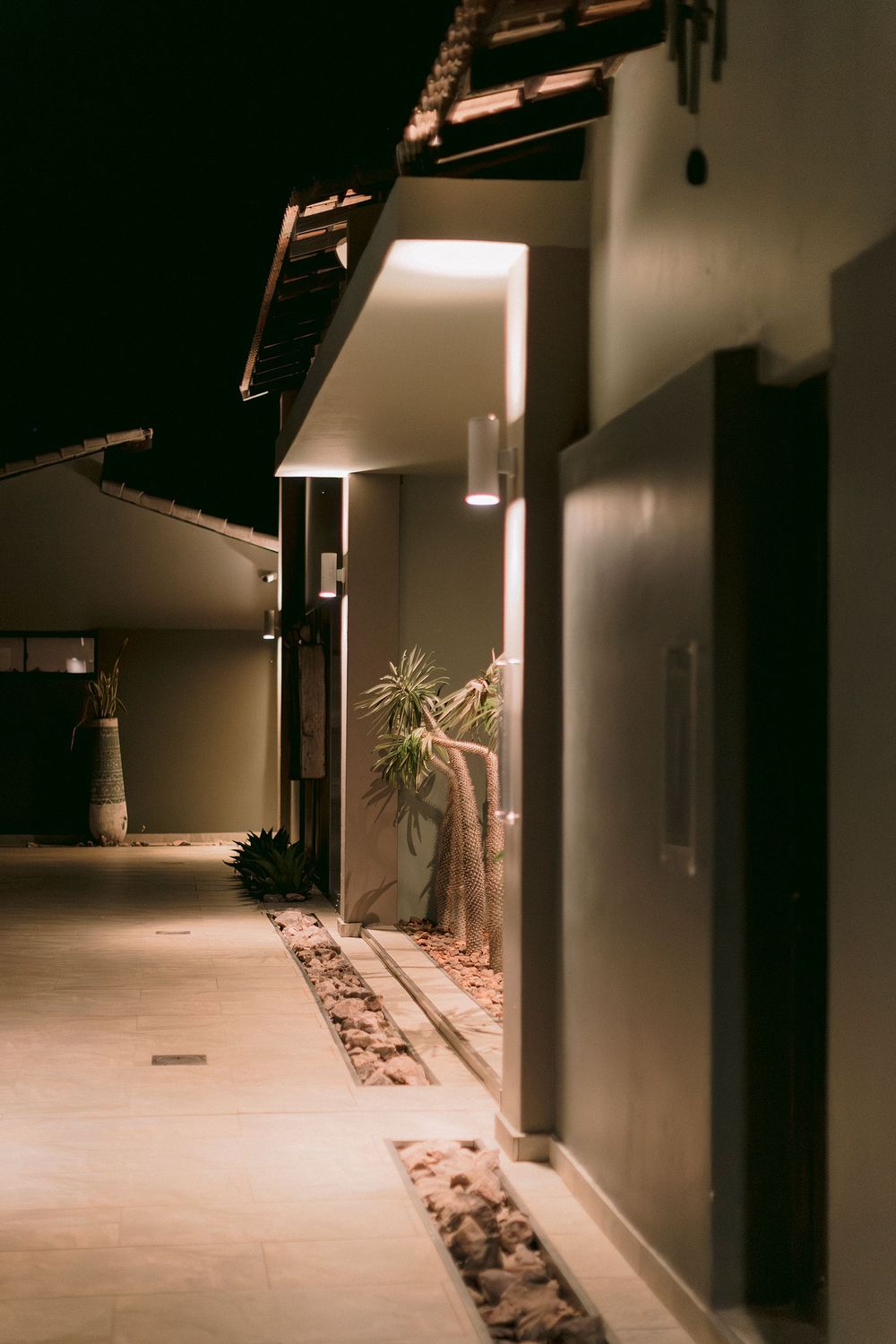 Illuminated modern building corridor at night with wall-mounted lights and tropical plants lining a stone pathway.