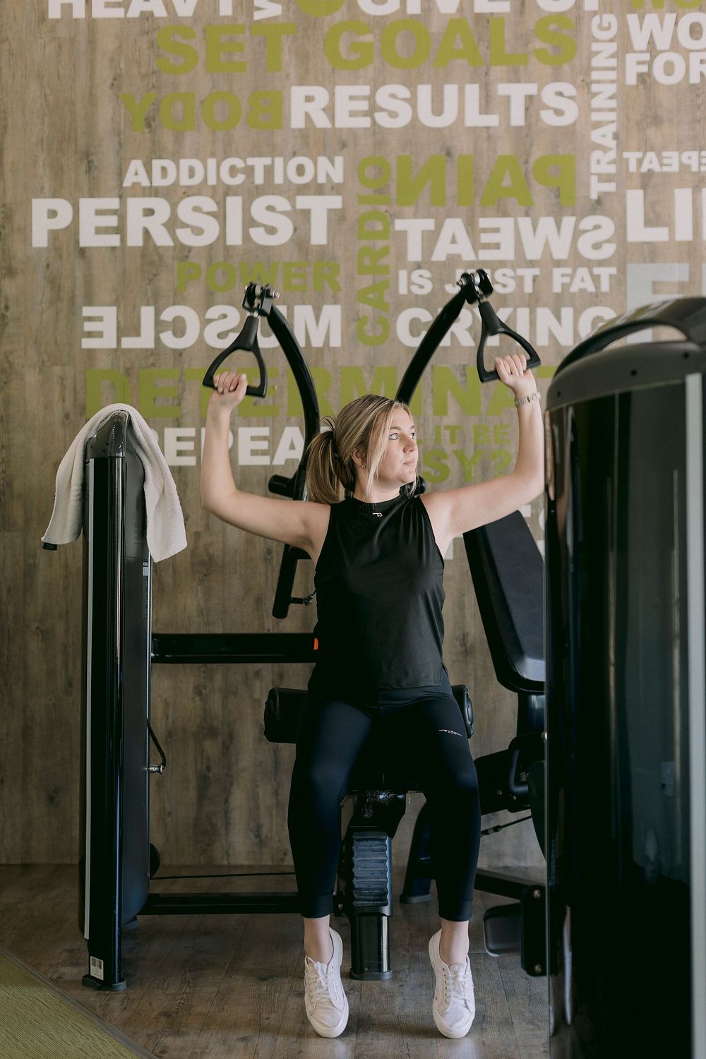 Woman in black workout clothes using a chest fly machine at a modern gym with motivational text on the wall.