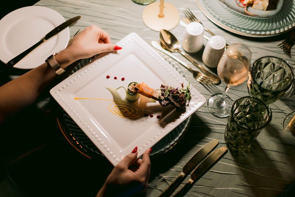 Woman with red nails dining at elegant table, plating a gourmet dessert on white square plate with wine glasses.