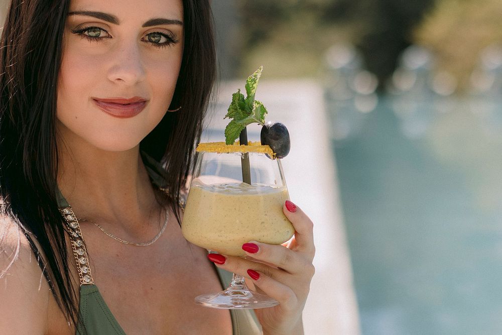 Close-up of woman with red nails holding a frothy yellow cocktail with mint garnish.