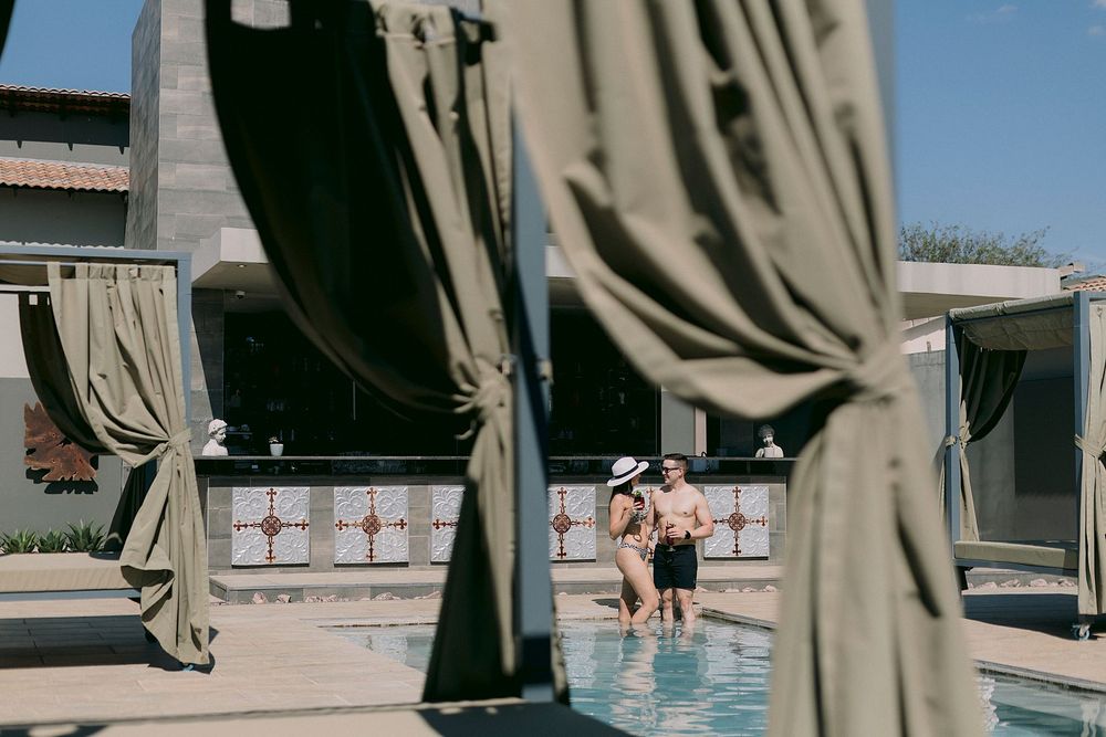 Couple standing together in a resort pool surrounded by elegant cabanas with olive green curtains on a sunny day.