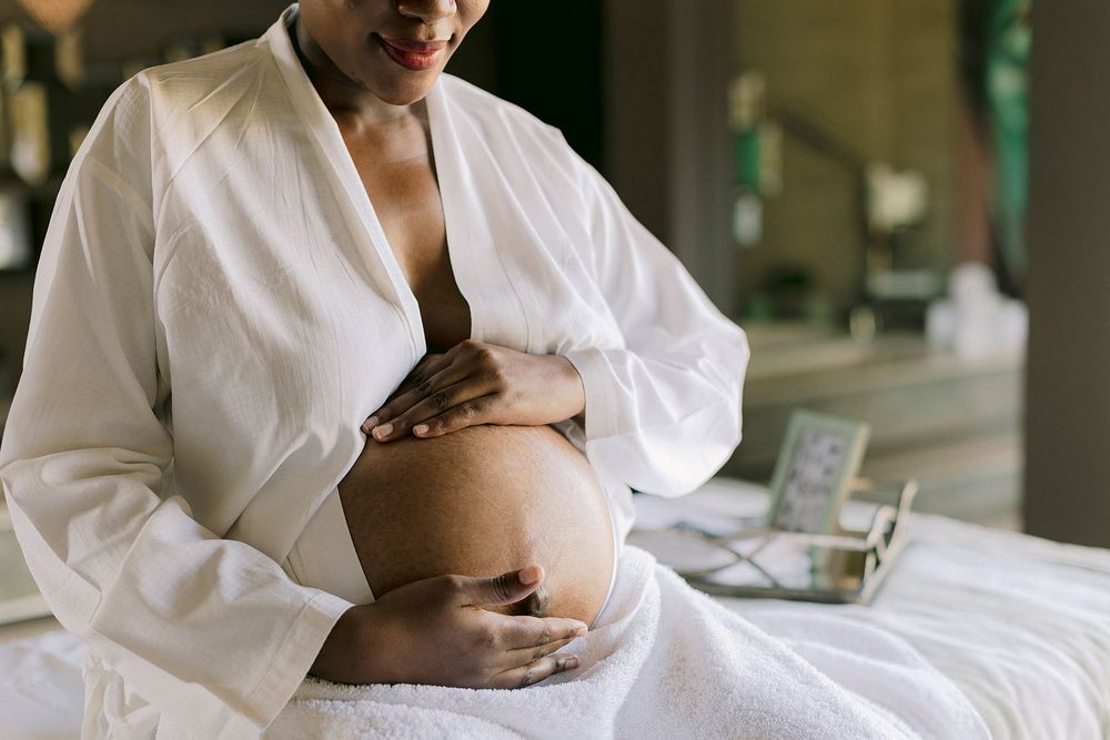 Pregnant woman in white robe sitting on bed cradling her baby bump during a peaceful morning routine.