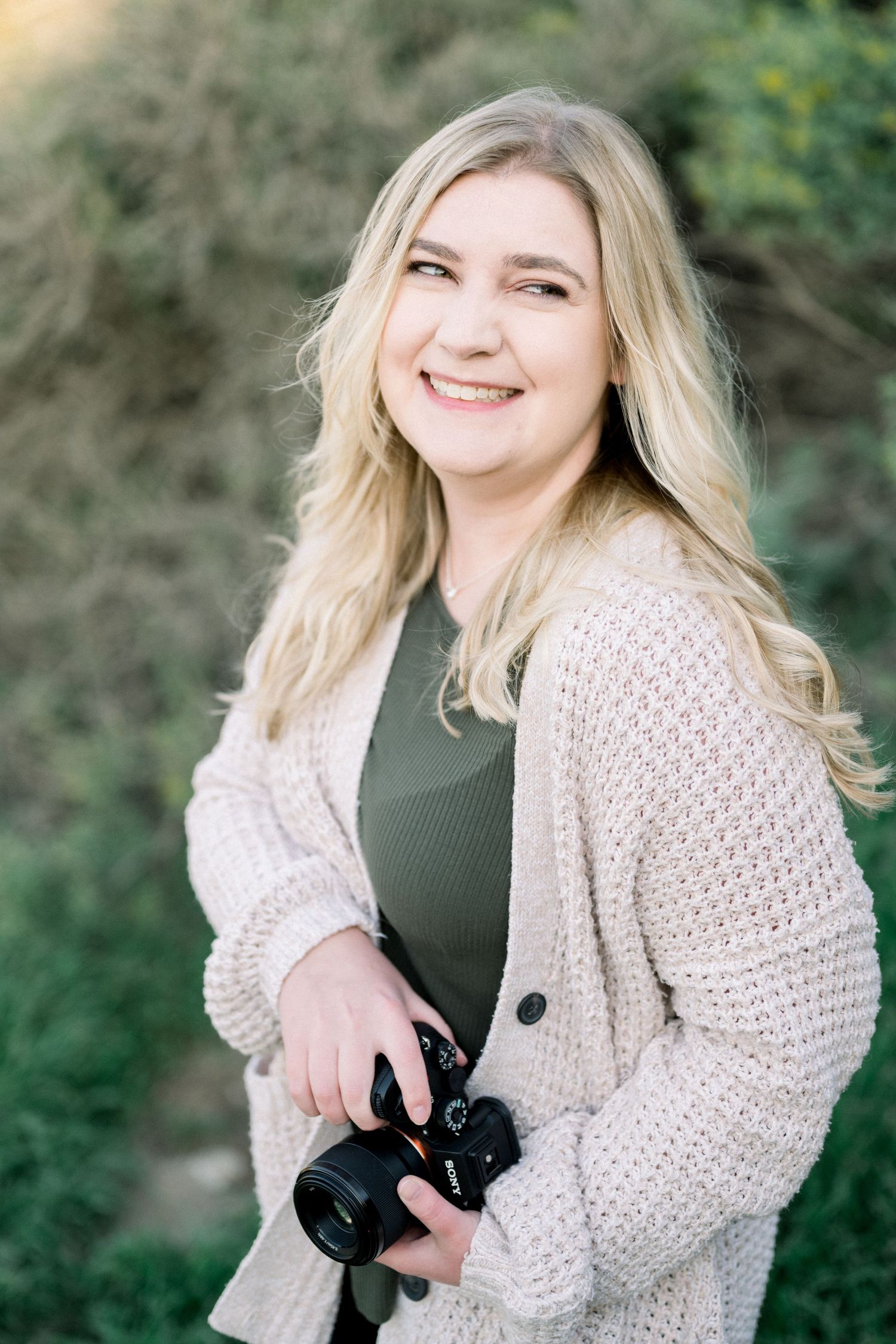 Smiling blonde woman holding a DSLR camera outdoors, wearing a cream cardigan and olive top.