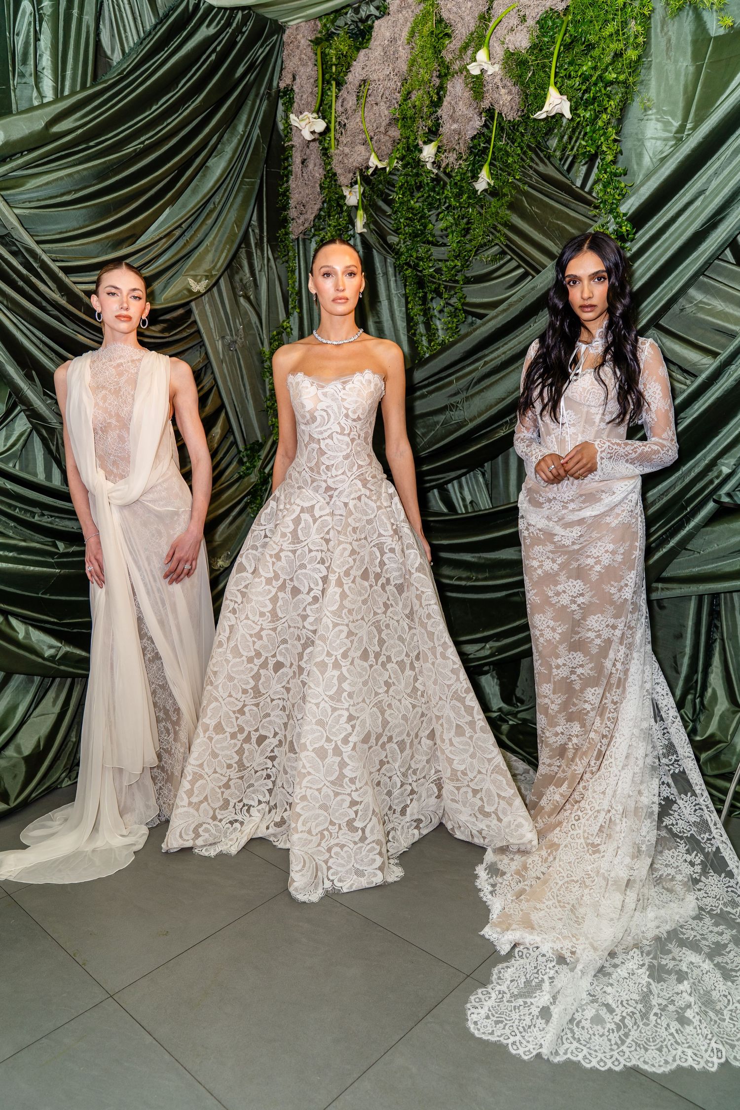 Three models showcase elegant white lace and sheer bridal gowns against a lush green botanical backdrop.