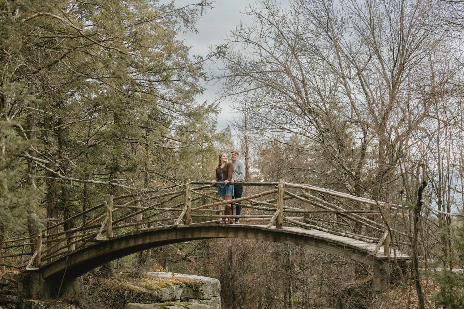Couple standing on a rustic wooden arched bridge surrounded by bare winter trees in a forest setting.