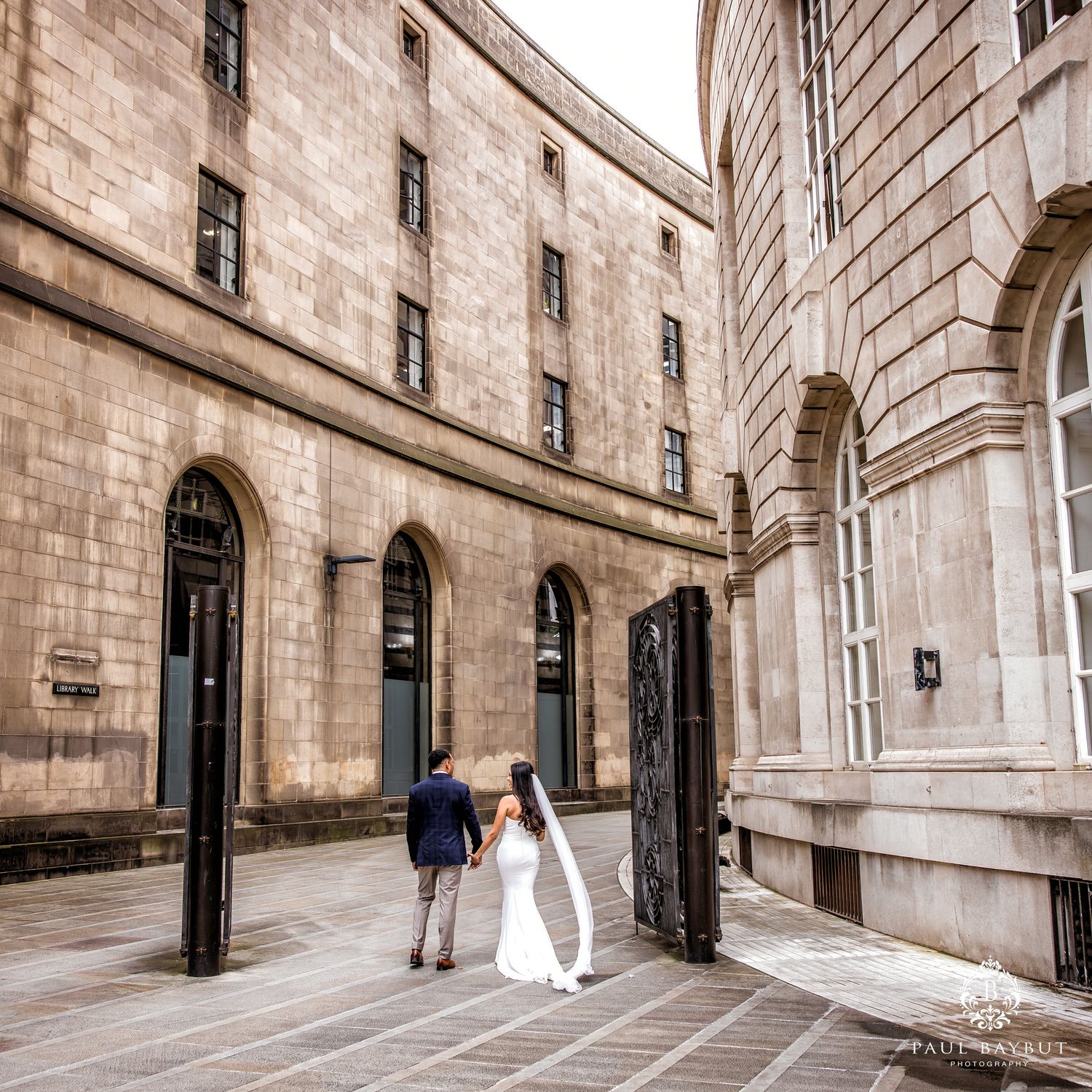 A bride and groom walk hand-in-hand through an elegant stone courtyard of a historic building.