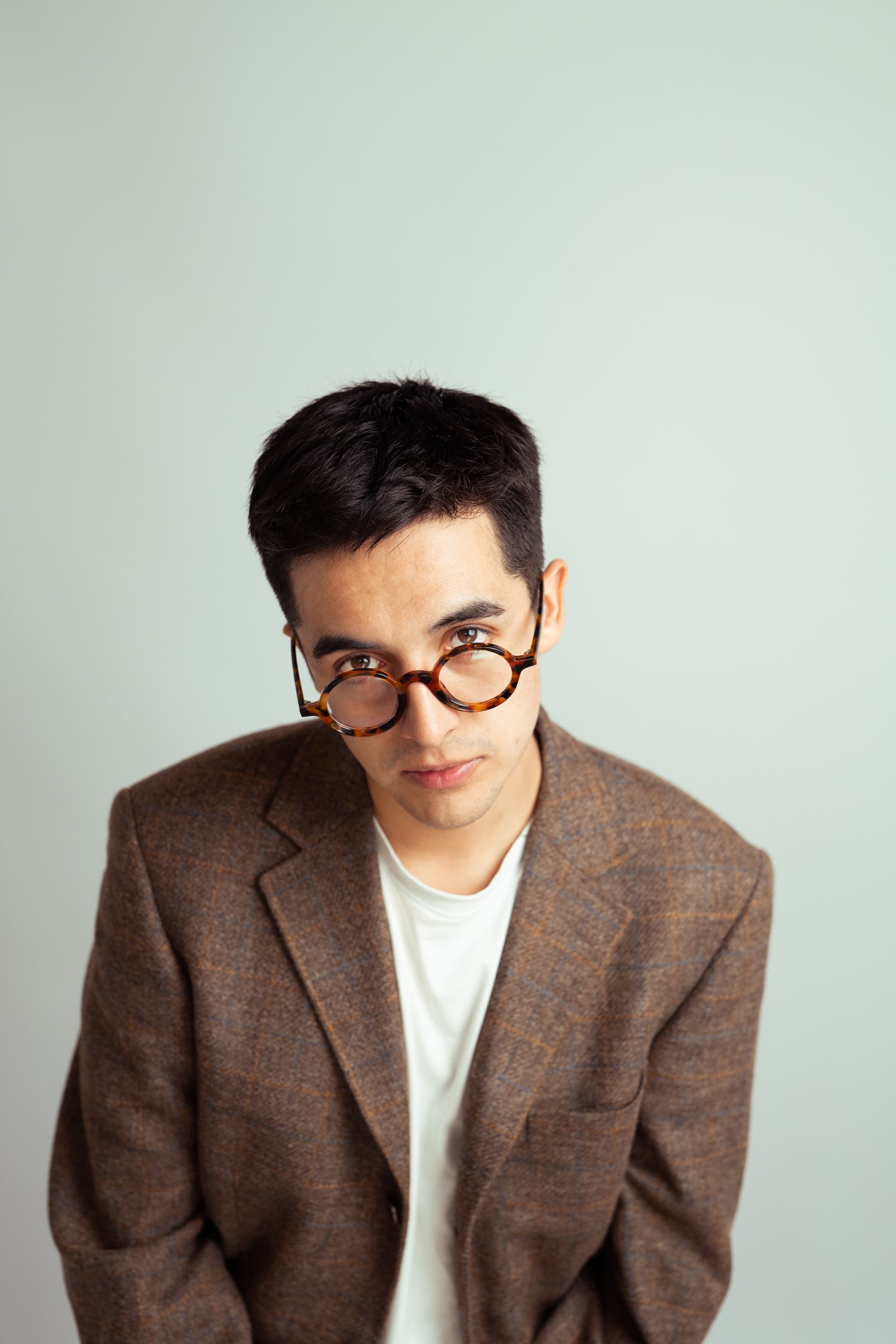 A young man in a brown tweed blazer and round tortoiseshell glasses poses against a light gray background.