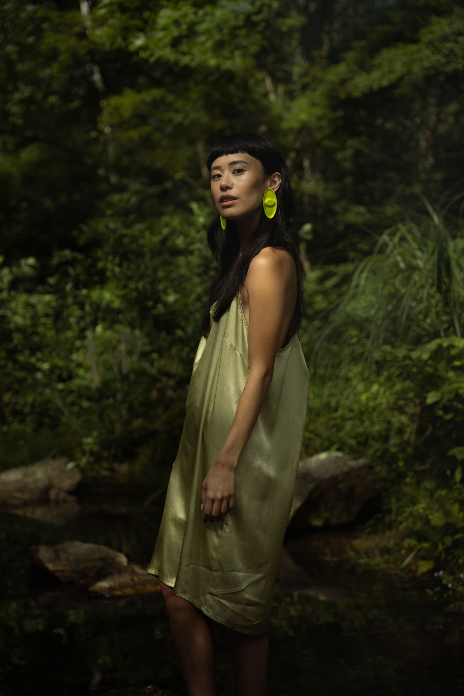 A woman in a sage green satin slip dress with yellow earrings stands in a lush forest setting.