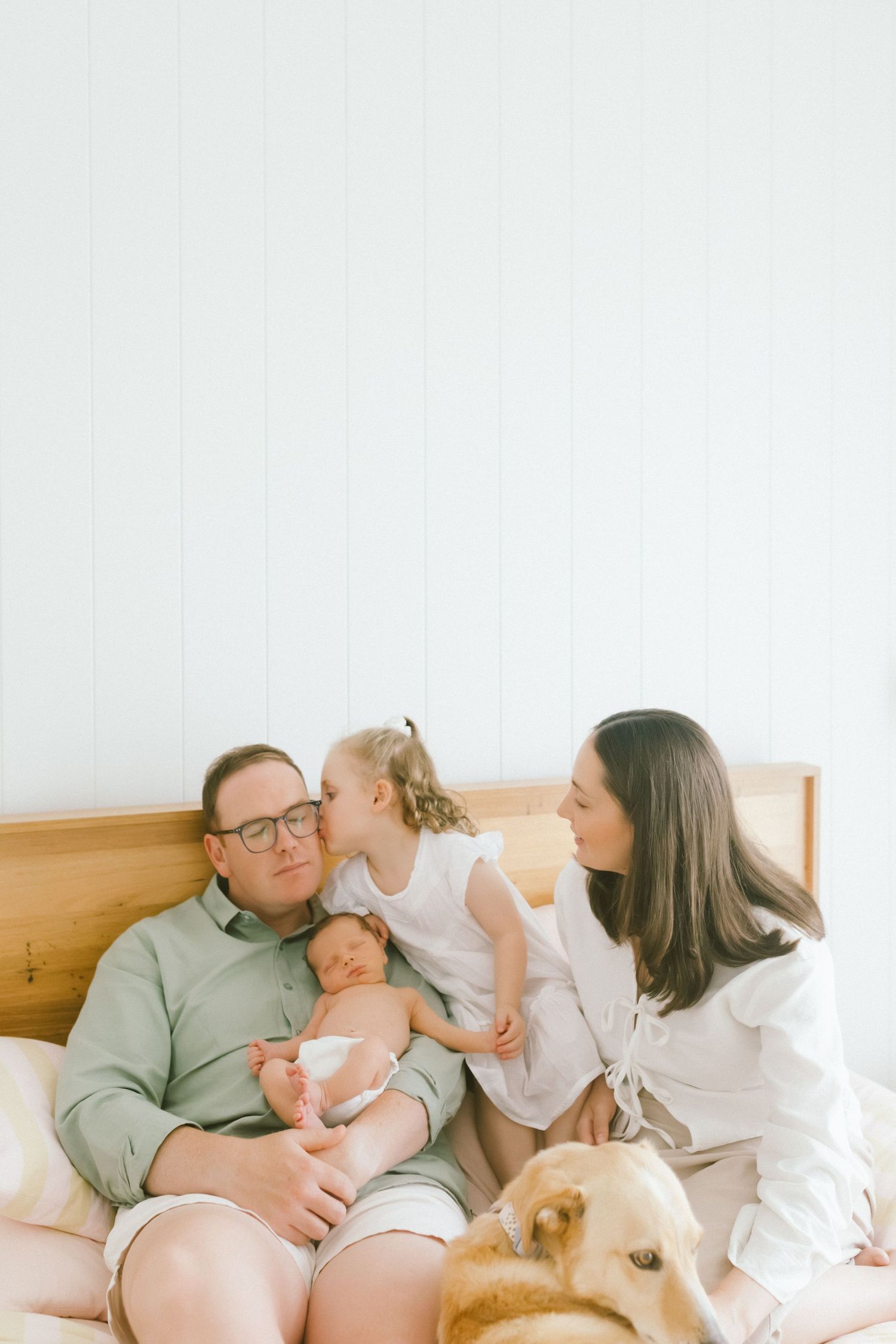 Family with newborn baby, toddler girl, and golden retriever dog sitting together on a bed in a bright room.