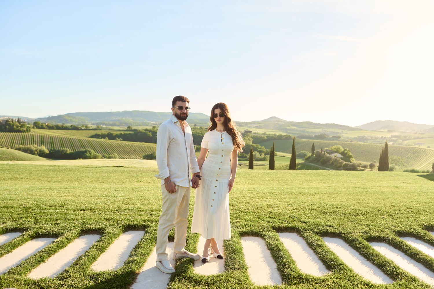 Couple in white attire holding hands in a scenic Tuscany vineyard landscape on a sunny day.