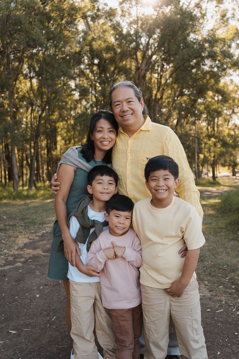 Happy Asian family of five smiling together outdoors in a sunny park surrounded by tall green trees. family photoshoot in rouse hill