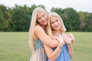 Two blonde teenage sisters embrace and smile warmly while standing together in a grassy outdoor field.