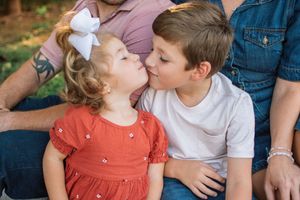A little girl in a red dress and a boy in a white shirt share a sweet kiss while sitting with family.