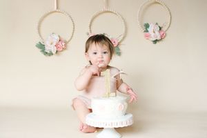 Adorable baby girl celebrating first birthday with a white cake in a soft pink studio setting.