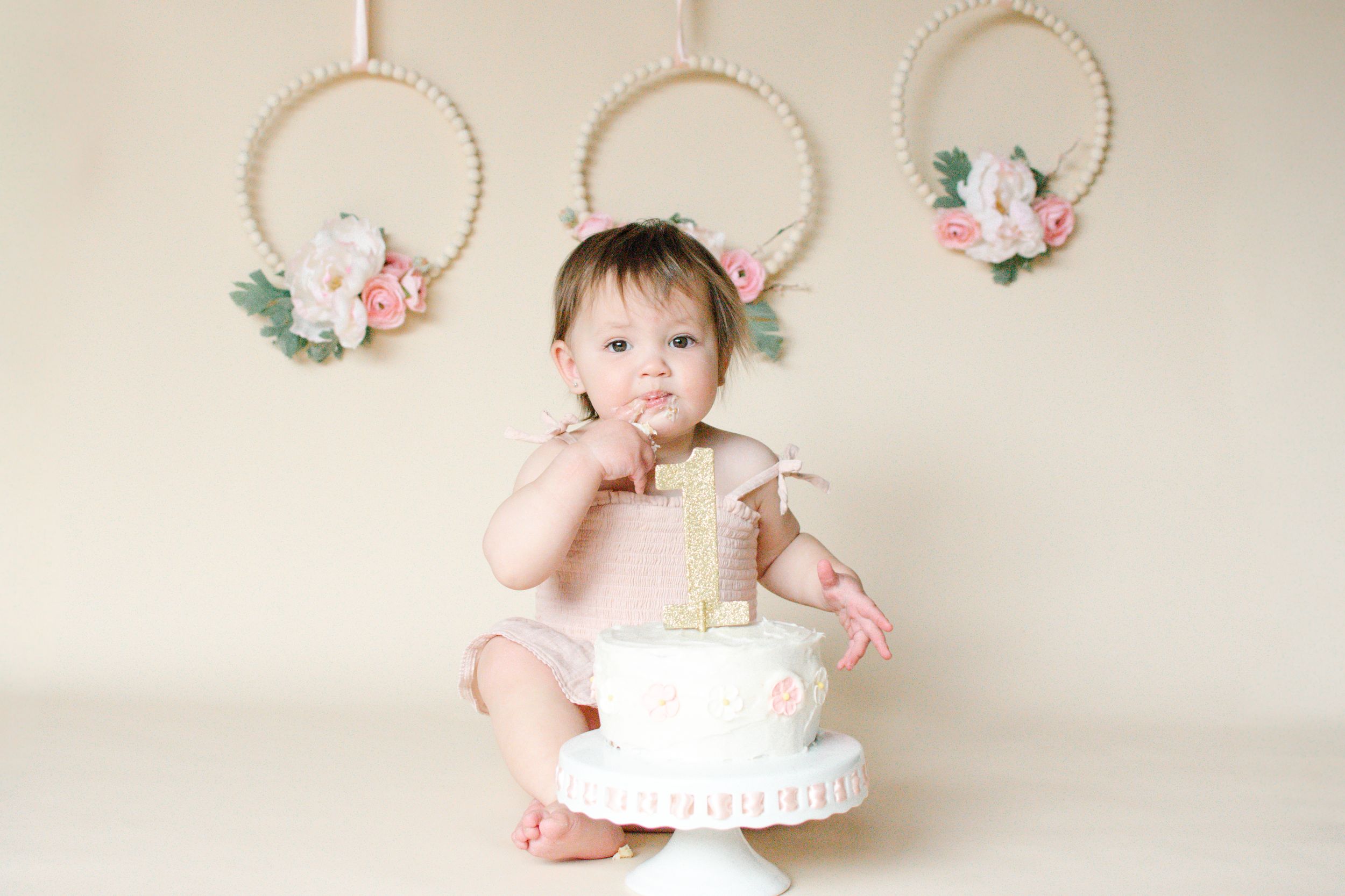 Adorable baby girl celebrating first birthday with a white cake in a soft pink studio setting.