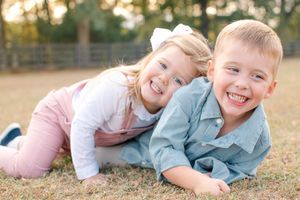 A cheerful young boy and girl laugh together while lying on grass in a sunny outdoor setting.