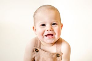 Smiling baby in brown overalls poses against a white background in a cute portrait photo.