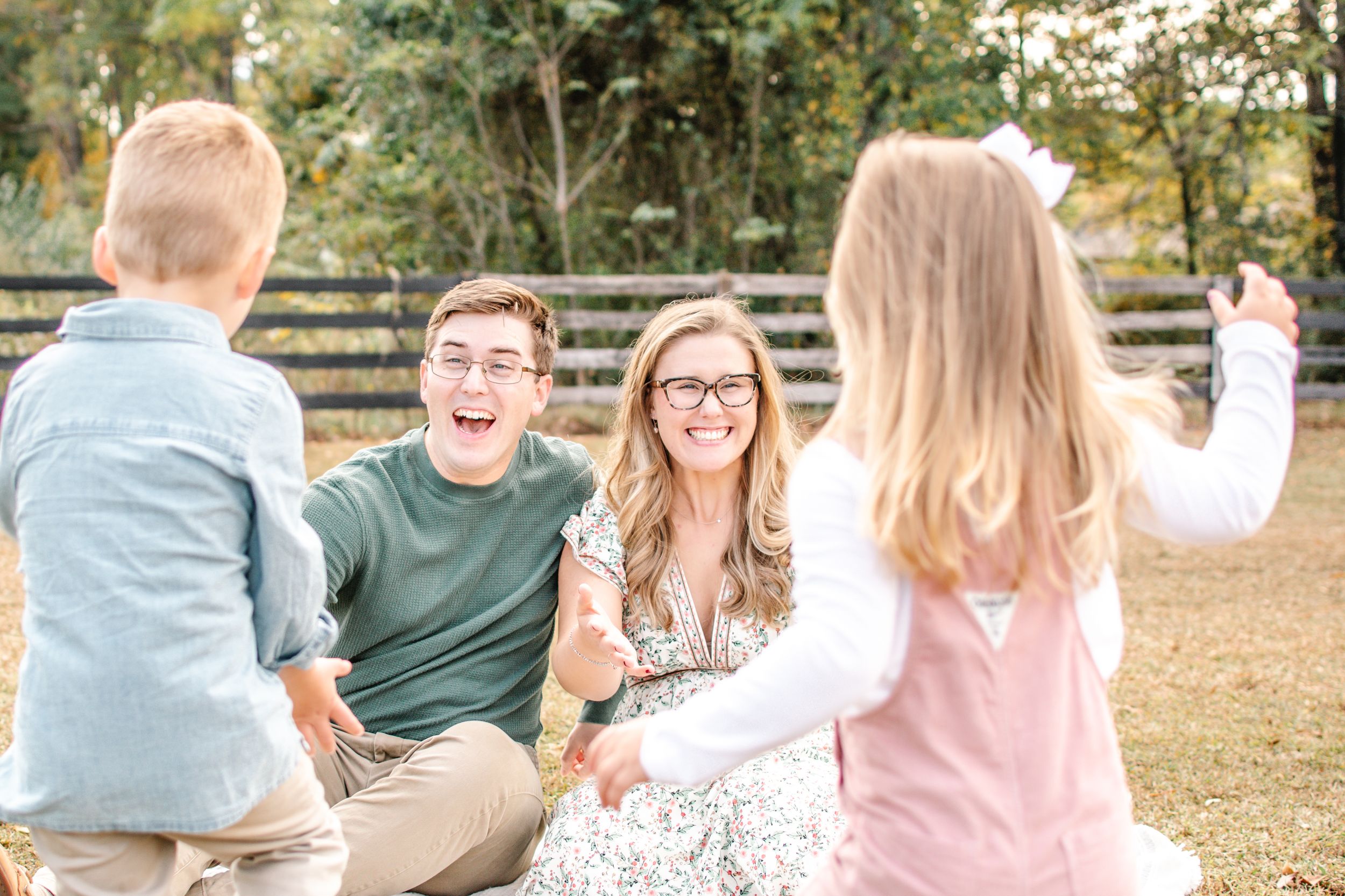 Two young children run toward their laughing parents who sit together on grass near a wooden fence.