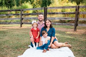 A happy family of four sits on a white blanket on grass near a wooden fence during golden hour.