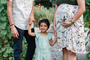 A young girl in a mint dress holds hands with her pregnant mother and father while walking in a lush garden.