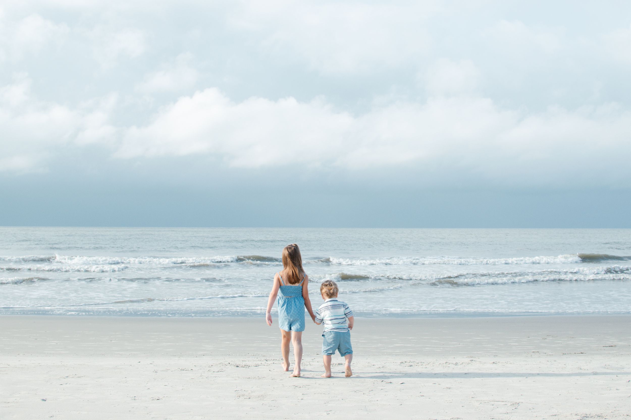 A mother and young child hold hands while standing on a sandy beach, gazing at the ocean waves under a cloudy sky.
