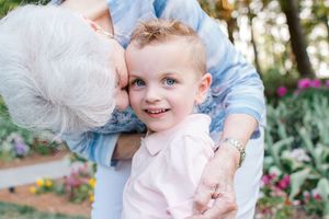 An elderly woman lovingly kisses a smiling blue-eyed toddler boy in a colorful garden setting.