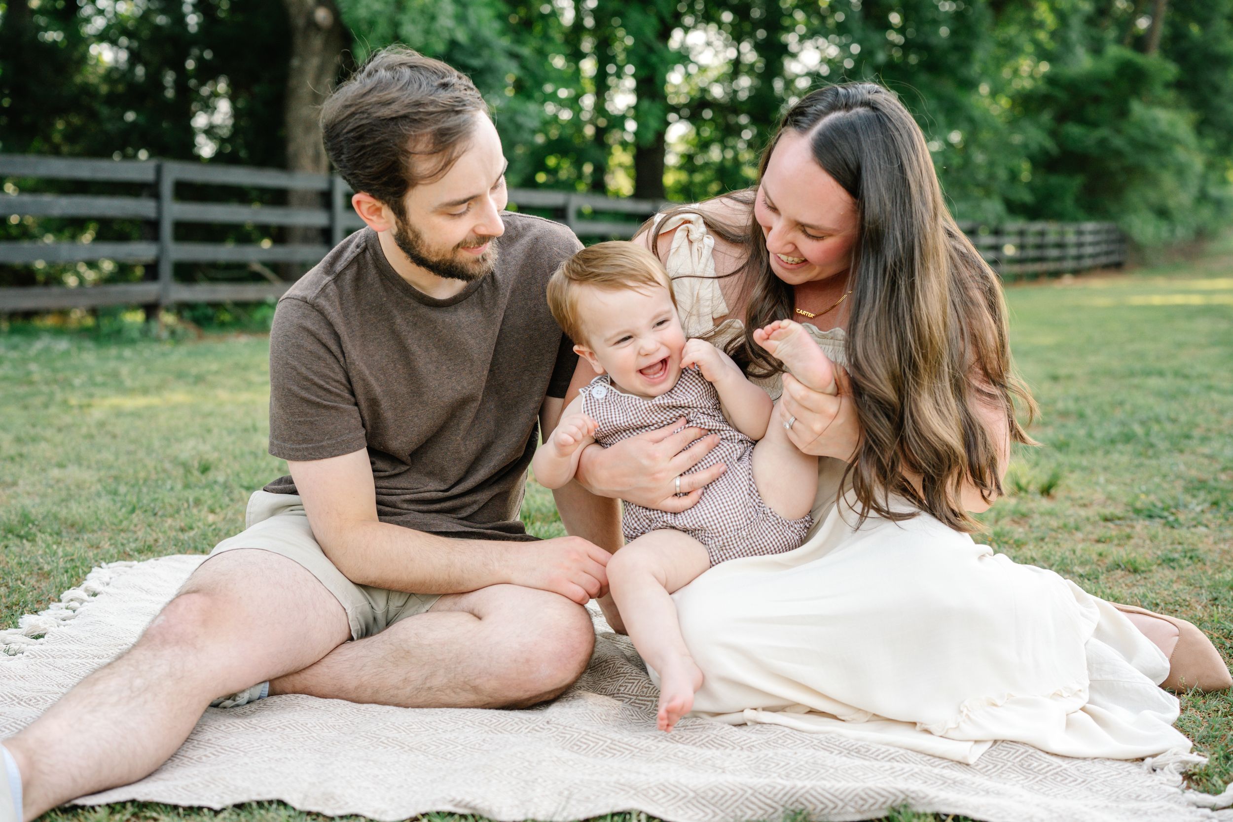 Happy family with parents and laughing baby sitting on blanket in green outdoor park setting.