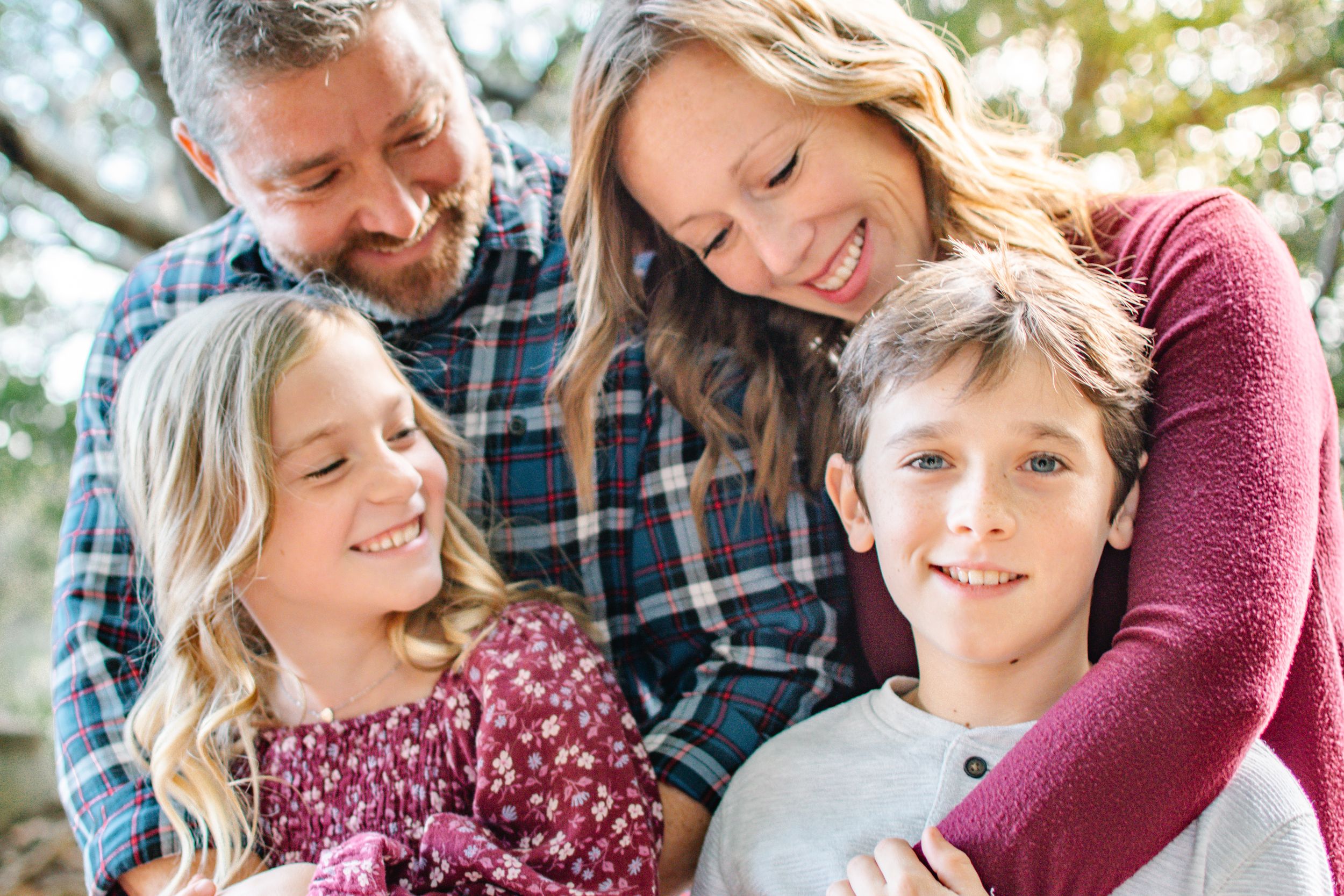 A happy family of four shares a warm embrace and laughter together under a shady tree outdoors.