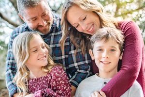 A happy family of four shares a warm embrace and laughter together under a shady tree outdoors.